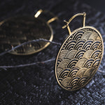 A close-up shot of two intricately designed SEIGAIHA Oversized Japanese Geometry Disc Earrings in Gold by Kaya Movement, each featuring a pattern of overlapping arcs. The unisex 16 gauge statement earrings are displayed on a dark textured surface, with one earring in sharp focus and the other slightly blurred in the background.