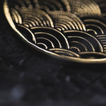 Close-up of the SEIGAIHA Oversized Japanese Geometry Disc Earrings in Gold by Kaya Movement, intricately engraved with traditional seigaiha wave designs reminiscent of tribal jewelry. The finely textured surface, set against a dark background, accentuates the golden engravings.