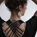 A person with wavy brown hair is seen from behind, showcasing intricate tattoos on their upper back and arm. They are wearing a black outfit with straps crossing at the back. Large round SEIGAIHA Oversized Japanese Geometry Disc Earrings in Silver by Kaya Movement are visible, and the background is white.