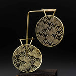 The SEIGAIHA Oversized Japanese Geometry Disc Earrings in Gold by Kaya Movement are displayed on a black background. Each earring showcases an intricate, repetitive semi-circular wave pattern, creating a visually appealing and symmetrical design. The earrings hang gracefully from a gold hook.