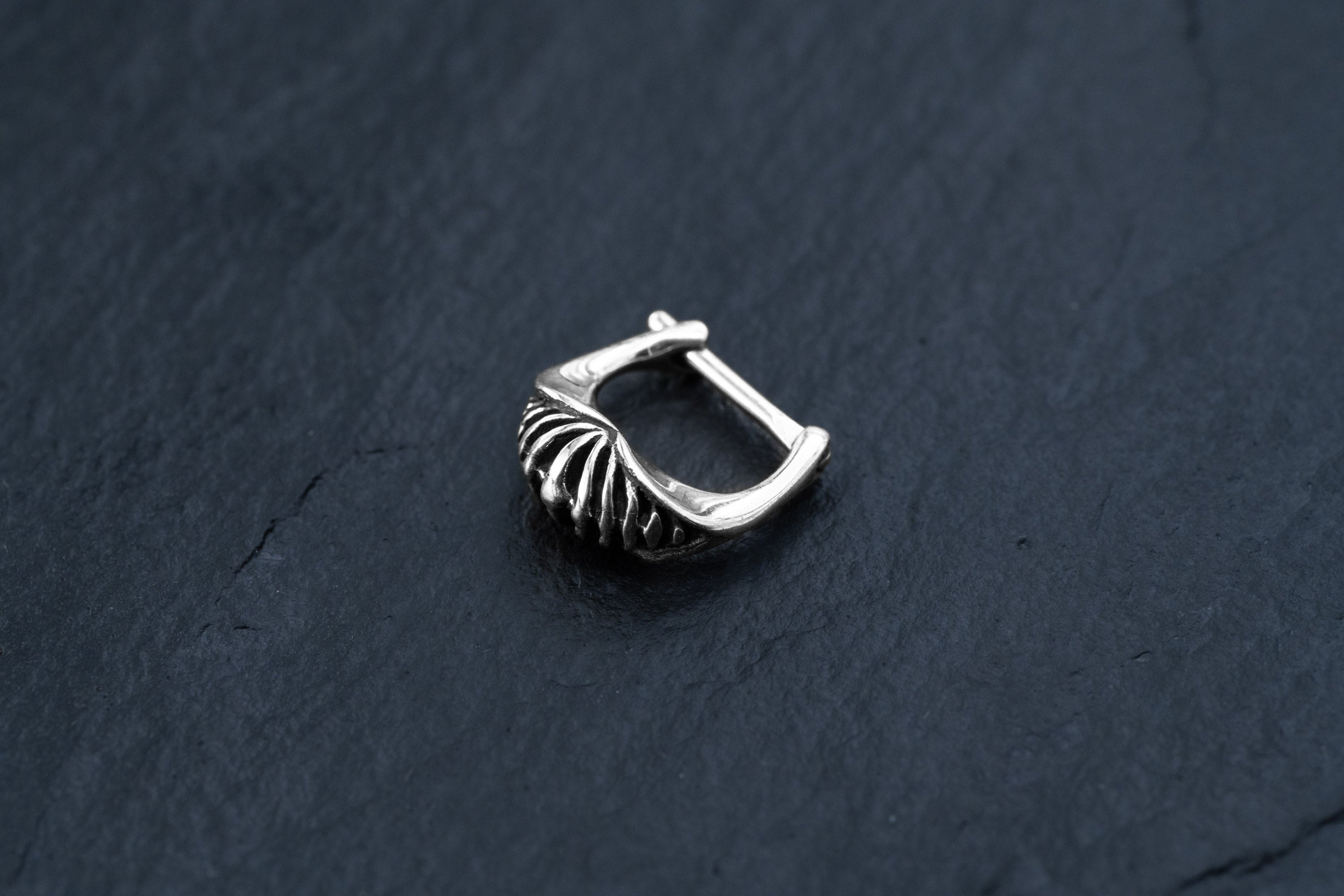 A close-up of a single DIARA Biomechanical Clicker Hoop Septum Nose Ring in Silver by Kaya Movement, with a textured, ridged design. The unisex 16 gauge earring is placed on a dark, slate-like surface, showcasing its intricate details.