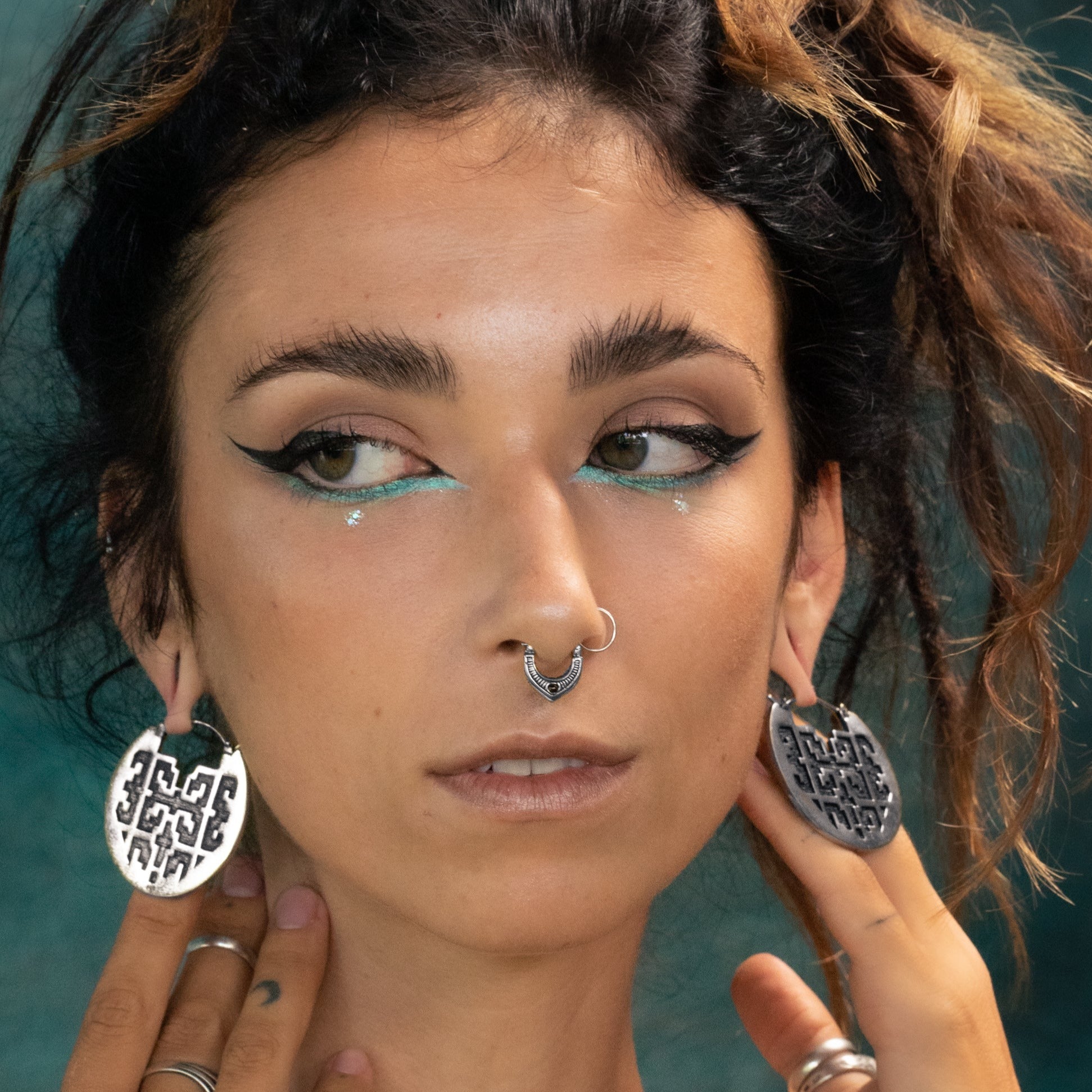 A person with long, dark hair featuring highlights wears bold winged eyeliner accented with turquoise, a nose ring, and large patterned hoop earrings. They hold their Kaya Movement CONIBOS Tribal Shipibo Hoop Disc Earrings in Silver (16 gauge) with both hands while gazing sideways. The background is a softly blurred teal color.