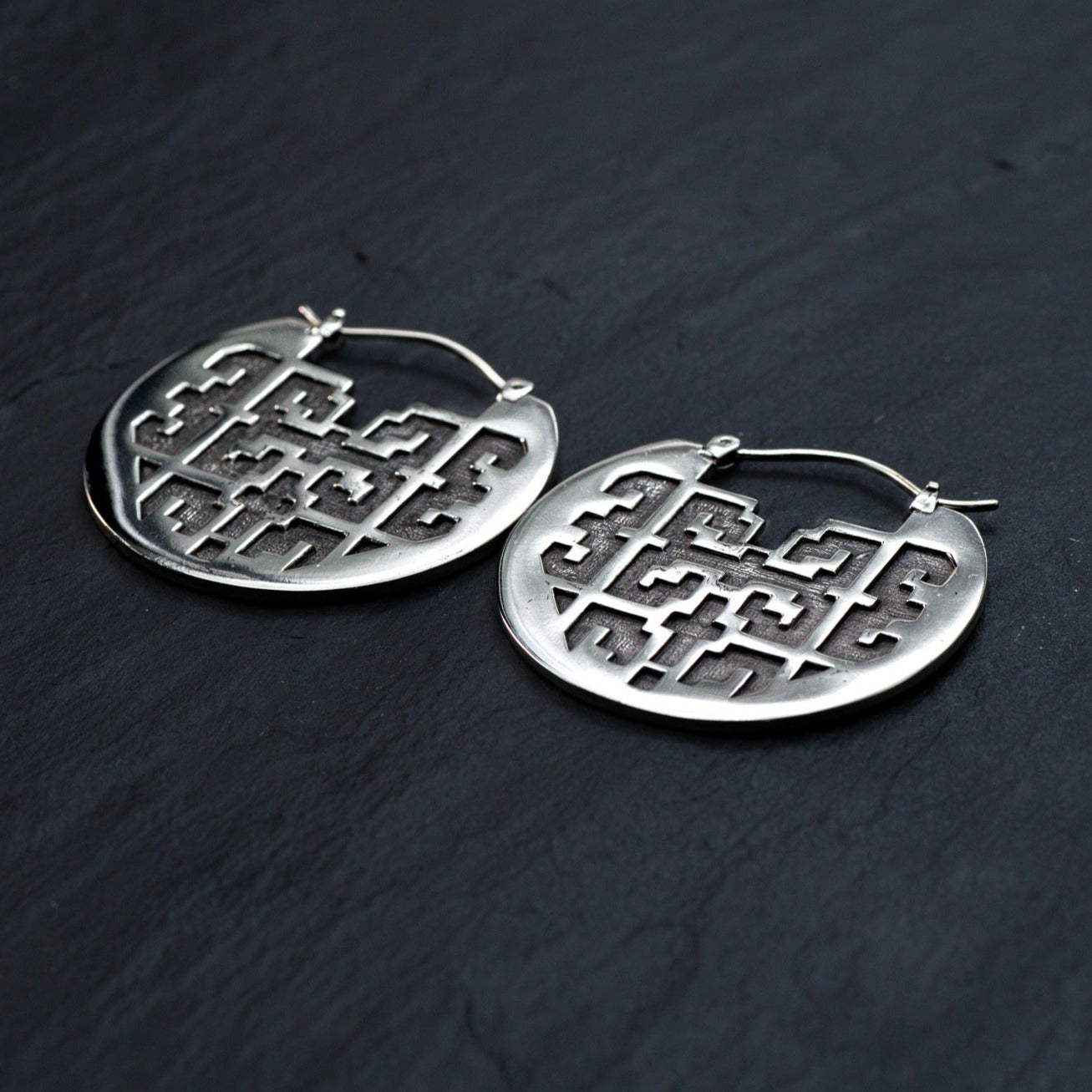 Introducing the CONIBOS Tribal Shipibo Hoop Disc Earrings in Silver by Kaya Movement: A pair of 16 gauge, round silver brass earrings with an intricate geometric cut-out design. Ideal for dark boho body jewelry enthusiasts, these tribal earrings are displayed on a dark, textured surface.