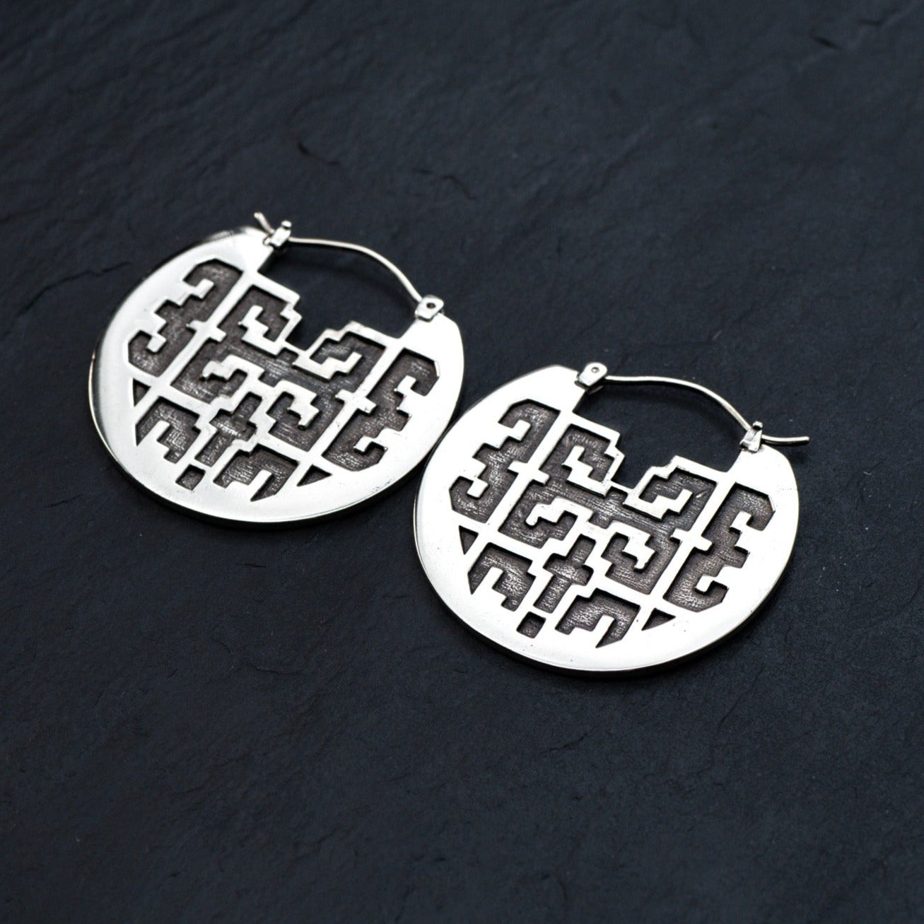 The CONIBOS Tribal Shipibo Hoop Disc Earrings in Silver by Kaya Movement, composed of silver brass, gracefully rest on a dark surface. These 16-gauge earrings feature intricate, geometrical cut-out designs that create patterns within circular shapes. Each earring is affixed to a hinged hoop closure. The overall look is elegant and modern, making them perfect for enthusiasts of dark boho body jewelry.