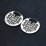 The CONIBOS Tribal Shipibo Hoop Disc Earrings in Silver by Kaya Movement, composed of silver brass, gracefully rest on a dark surface. These 16-gauge earrings feature intricate, geometrical cut-out designs that create patterns within circular shapes. Each earring is affixed to a hinged hoop closure. The overall look is elegant and modern, making them perfect for enthusiasts of dark boho body jewelry.