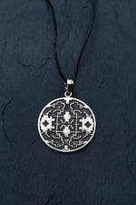 A round, white brass pendant from Kaya Movement, the ICARO - White Brass Handmade Shipibo Icaros Pendant, hangs from a thin black cord. The pendant features intricate geometric patterns with oxidized details set against a dark textured background.