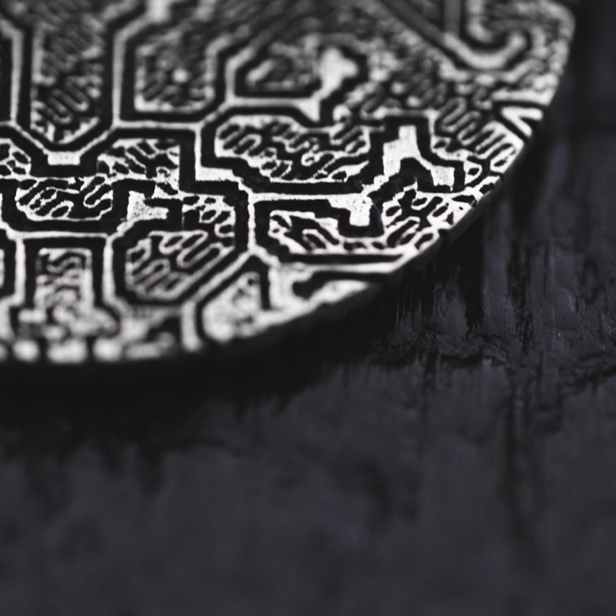 A close-up of the OLIVIA Tribal Shipibo Seamless Disc Hoop Earrings in Silver, designed by Kaya Movement, shows intricate raised geometric patterns on a dark, textured surface. The sharp lines and angles of the complex design are reminiscent of tribal art. The dark background contrasts sharply with the metallic surface of these 12-gauge earrings.