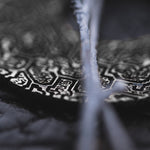 A close-up shot showcases the OLIVIA Tribal Shipibo Seamless Disc Hoop Earrings in Silver by Kaya Movement, highlighting the intricate geometric carvings against a dark, blurred background. The delicate and detailed patterns of these 12 gauge earrings are prominently featured, with a thin, out-of-focus element traversing the image diagonally.