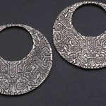 Close-up of two round, intricately patterned OLIVIA Tribal Shipibo Seamless Disc Hoop Earrings in Silver by Kaya Movement. The silver metal earrings feature geometric designs and have an open center, creating a crescent moon shape. The plain dark background accentuates the detailed textures of the 12-gauge earrings.