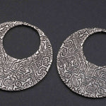 A close-up image of the OLIVIA Tribal Shipibo Seamless Disc Hoop Earrings in Silver by Kaya Movement, showcasing their intricately designed, circular shape with a textured, abstract pattern. These 12 gauge boho statement earrings feature a large central hole that gives them a hoop-like appearance and are set against a plain dark background.