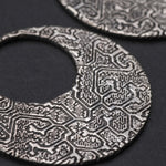 Close-up of the OLIVIA Tribal Shipibo Seamless Disc Hoop Earrings in Silver by Kaya Movement. These 12-gauge, round, textured metal pendants feature intricate abstract patterns and resemble boho statement earrings. With an open center like hoops and set against a dark background, the design is complex, showcasing raised geometric shapes and lines.