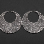 A pair of OLIVIA Tribal Shipibo Seamless Disc Hoop Earrings from Kaya Movement lies on a dark background. These intricately engraved, circular silver earrings feature detailed geometric patterns and textures, lending an antique, handcrafted appearance to the boho statement piece. The 12 gauge earrings are open-hoop in style.