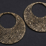 A pair of large, round, gold OLIVIA Tribal Shipibo Seamless Disc Hoop Earrings in 12 gauge by Kaya Movement on a dark background. Each earring features intricate, abstract patterns engraved onto the surface, giving them a textured and ornate appearance. The patterns resemble interconnected geometric shapes and lines, making them stunning pieces of tribal body jewelry.