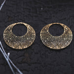 The OLIVIA Tribal Shipibo Seamless Disc Hoop Earrings in Gold by Kaya Movement are elegantly showcased on a dark, textured surface. These 12 gauge earrings feature intricately designed, circular gold brass hoops with geometric patterns and large cut-out centers that create a crescent-like shape. Their detailed craftsmanship gives them an antique, artisanal appearance reminiscent of tribal body jewelry.