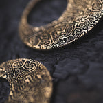 Close-up image of the OLIVIA Tribal Shipibo Seamless Disc Hoop Earrings in Gold by Kaya Movement, showcasing their intricate, textured patterns. The 12 gauge gold brass earrings are resting on a dark, rough surface, creating a striking contrast between the ornate design and the dark background. The shallow focus emphasizes the exquisite details of these elegant accessories.