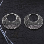 Placed on a dark textured surface, the OLIVIA Tribal Shipibo Seamless Disc Hoop Earrings in Silver from Kaya Movement exude a striking and artistic appearance with their detailed and symmetrical abstract design. These 12 gauge boho statement earrings showcase intricate engraving, while thin strands of white material are visible on the left side.