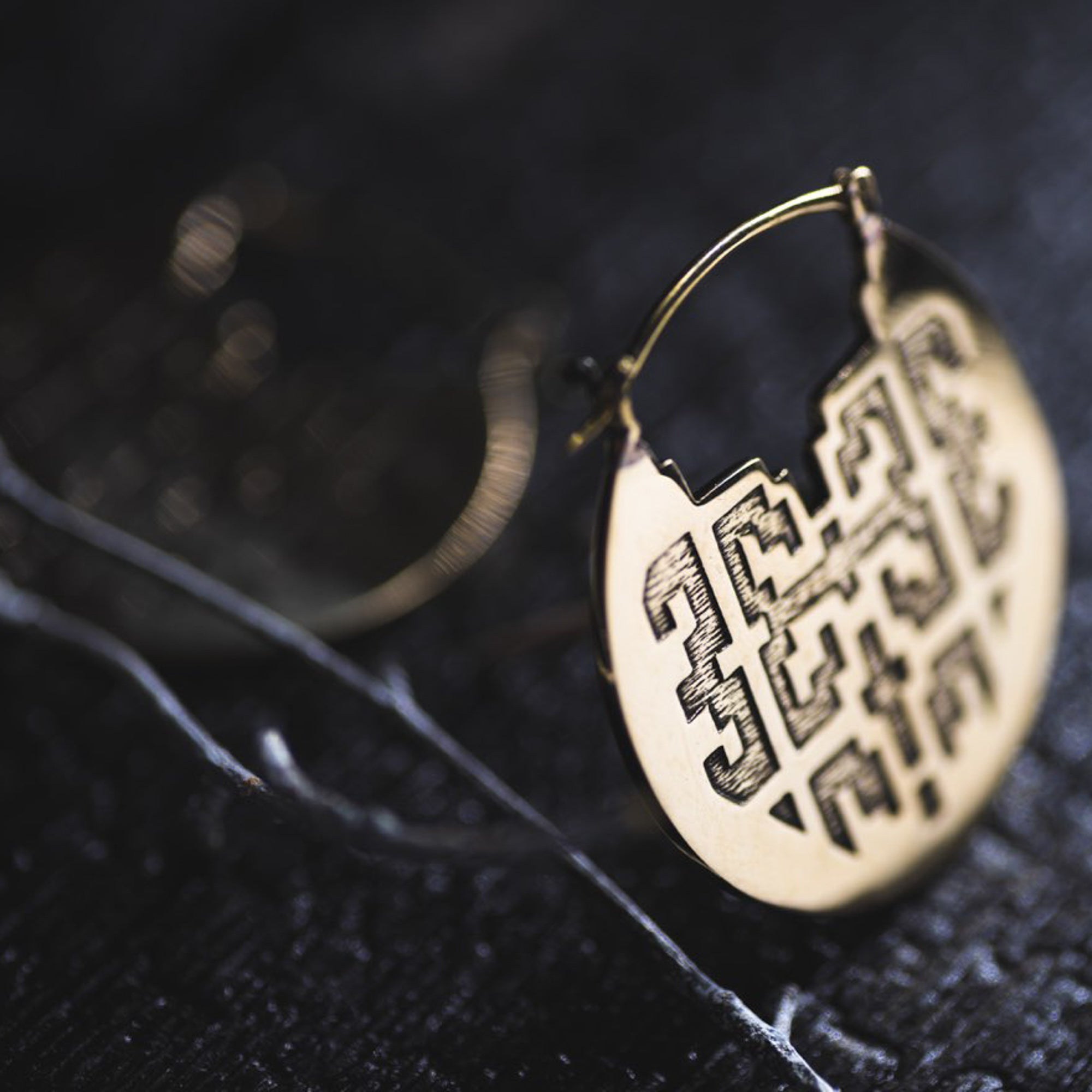 A close-up image showcases the intricately designed CONIBOS Tribal Shipibo Hoop Disc Earrings in Gold by Kaya Movement, resting on a dark surface. The earring features geometric cut-out patterns with a thin, curved wire forming the hoop part of the 16-gauge earring. A blurred second tribal earring can be seen in the background.