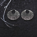 A pair of KONI Tribal Shipibo Endless Hoop Disc Earrings in Silver | 18 gauge by Kaya Movement lie on a textured, dark surface. The circular pattern on the intricate 925 sterling silver earrings evokes ancient or tribal body jewelry designs. A thin branch extends across the upper left corner, adding a natural element to the composition.