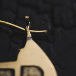 Close-up of a Kaya Movement CONIBOS Tribal Shipibo Hoop Disc Earring in gold on a dark background. This piece of jewelry features a hook-like attachment and part of a rectangular design with engraved or embossed lines. The polished, shiny surface perfectly embodies boho body jewelry aesthetics.