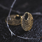 Close-up of two round, gold-colored KONI Tribal Shipibo Endless Hoop Disc Earrings in Gold (18 gauge) by Kaya Movement, intricately detailed with a boho design and placed on a dark textured surface. A thin branch-like object is partially visible in the foreground, adding a natural element to the composition.