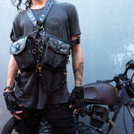 A person stands in front of a motorcycle, wearing a dark t-shirt with a logo, black pants, and fingerless gloves. An intricate SHIPIBO Tribal Apocalyptic Leather Belt & Shoulder Bag by Kaya Movement, featuring multiple pouches and metal buckles, complements their wasteland hip bag. The person's arms reveal visible tattoo art indicative of dystopian festivals.