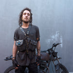 A person with long hair stands in front of an old, rustic motorcycle. They are wearing a dark t-shirt, Kaya Movement's SHIPIBO Tribal Apocalyptic Leather Belt & Shoulder Bag | Labradorite, harness with pouches, fingerless gloves, and have tattoos on their arm. The background is a plain gray wall.