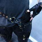 A person wearing black tactical pants and a black shirt is shown from the waist down. They sport the SHIPIBO Tribal Apocalyptic Leather Belt & Shoulder Bag by Kaya Movement, featuring multiple pockets and metal rings, reminiscent of dystopian festivals. One hand, covered by a fingerless glove, is tucking into a pocket of the bag attached to the belt adorned with Labradorite accents.