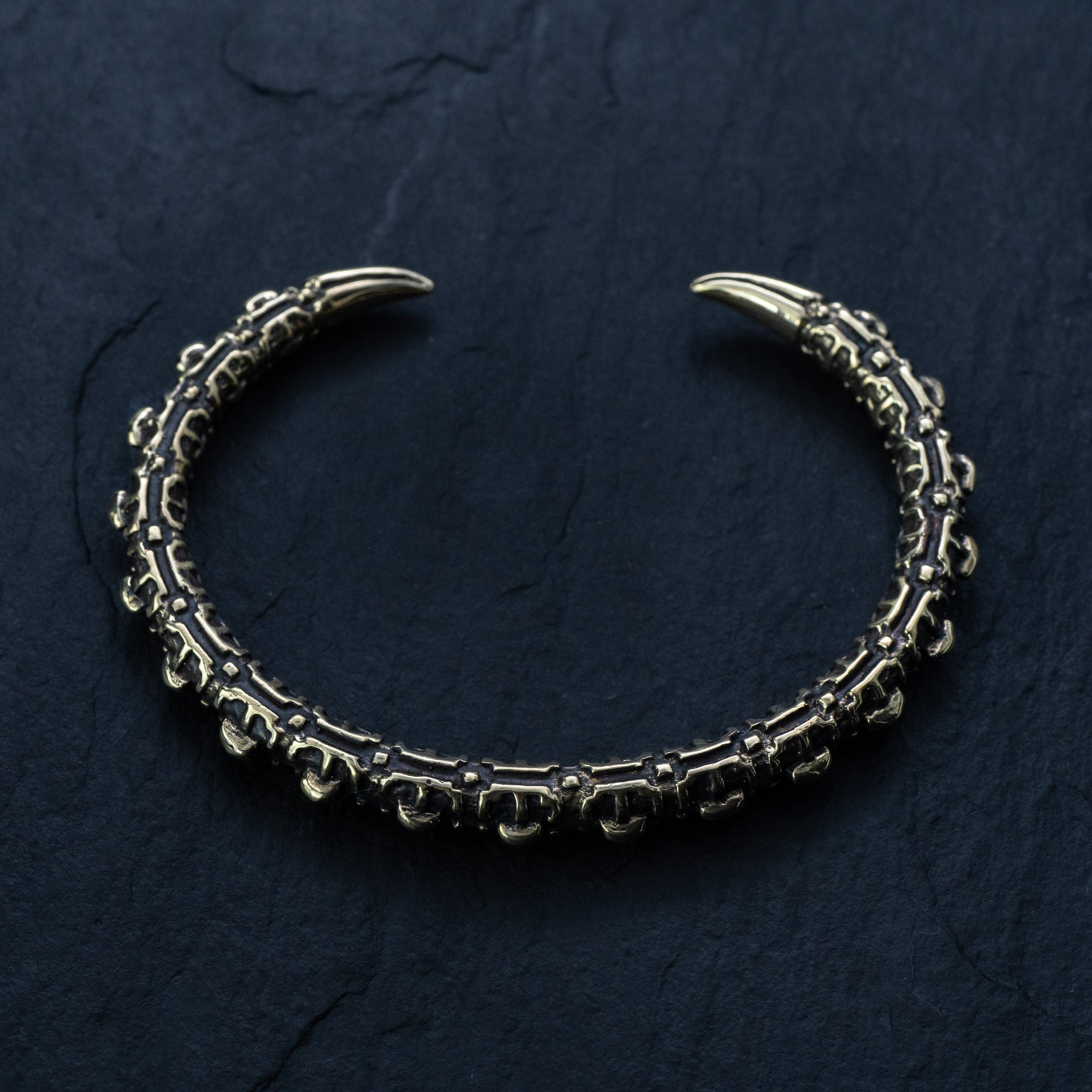 A gold, intricately designed adjustable cuff from Kaya Movement's SHIRO collection on a dark, textured background. The cuff is open-ended with two claw-like tips and features detailed, ornate patterns along its length, giving it a gothic and antique appearance reminiscent of a reptile spine design.
