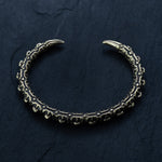 A gold, intricately designed adjustable cuff from Kaya Movement's SHIRO collection on a dark, textured background. The cuff is open-ended with two claw-like tips and features detailed, ornate patterns along its length, giving it a gothic and antique appearance reminiscent of a reptile spine design.
