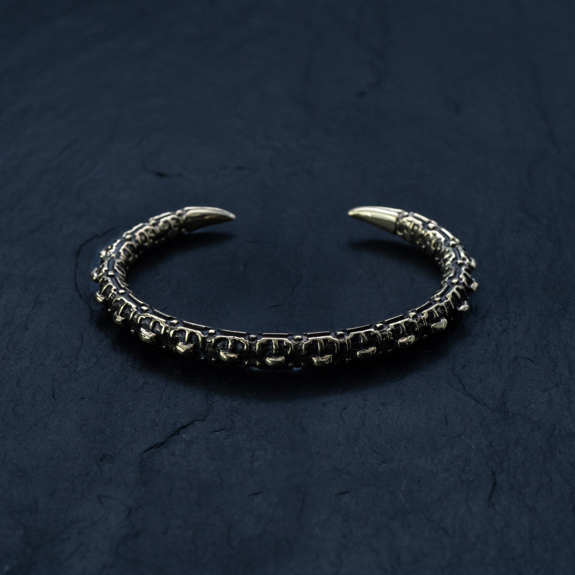 The SHIRO Men's Gold Cuff Bracelet from Kaya Movement, a rugged and intricately designed piece with a dark patina finish, rests on a dark textured surface. This adjustable cuff features claw-like tips and a reptile spine design, giving it an antique and warrior-like appearance that is perfect for those seeking a unique men’s gold bracelet.