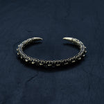 The SHIRO Men's Gold Cuff Bracelet from Kaya Movement, a rugged and intricately designed piece with a dark patina finish, rests on a dark textured surface. This adjustable cuff features claw-like tips and a reptile spine design, giving it an antique and warrior-like appearance that is perfect for those seeking a unique men’s gold bracelet.