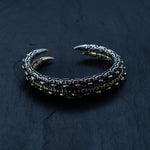 The SHIRO Bangle in Silver by Kaya Movement is a men's cuff bracelet inspired by an animal's talon, with detailed, scaled accents around the band. Crafted in oxidized silver, this unique piece rests on a dark, textured surface.