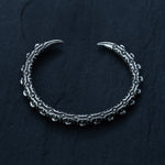 The SHIRO Bangle in Silver from Kaya Movement, an intricately designed men's cuff bracelet in oxidized silver with a rugged, industrial look, lies on a dark textured surface. The bracelet features an elaborate pattern resembling interconnected gears and has pointed ends that curve slightly upward.