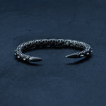 The SHIRO Bangle in Silver by Kaya Movement is displayed in a semi-circle on a textured dark surface. This men's cuff bracelet, made from oxidized silver, features a segmented, chain-like design with pointed ends. Its reflective and intricate appearance highlights its robust construction and fine details.