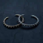 Two intricately designed, claw-shaped men's cuff bracelets lie on a dark textured surface. One bracelet is the SHIRO Bangle in oxidized silver by Kaya Movement, and the other is gold. Both feature a rugged, segmented design with sharp, pointed ends resembling talons and subtle Shipibo patterns woven into their form.