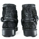 Introducing the Kaya Movement MAGMA Wasteland Studded Leather Ankle Combat Boots, a pair of rugged black punk rock boots showcasing intricate stitching, textured leather, and metallic studs. From the back, you'll notice decorative straps with buckles and thick, sturdy soles that beautifully complement their distressed finish for a stylishly worn look.