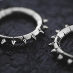 Close-up image of the KALASH Medium Spike Hoop Clicker Ear Weights in Silver Brass by Kaya Movement, arranged on a dark textured surface. The metallic shine of the 4 gauge ear weights contrasts against the background, highlighting their intricate detailing and unique style reminiscent of tribal punk industrial jewelry.