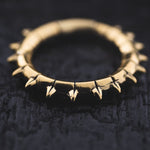 A detailed shot captures the KALASH Medium Spike Hoop Clicker Ear Weights in Gold Brass by Kaya Movement, showcasing their intricate design with evenly spaced outward-pointing spikes. The gold brass material shines brilliantly against the dark, textured background, enhancing the rich and detailed look of these 4-gauge earrings.