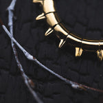 Close-up of a KALASH Medium Spike Hoop Clicker Ear Weight in Gold Brass from Kaya Movement, partially visible, placed on a textured black surface. Thin white and brown branches in the foreground add contrast to the dark background and metallic ear weight, reminiscent of earrings designed for stretched ear lobes.