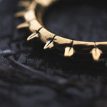 Close-up image of Kaya Movement's KALASH Medium Spike Hoop Clicker Ear Weights in Gold Brass, designed for 4 gauge ears, resting on a dark textured surface. The polished spikes are evenly spaced around the circular design, casting small reflections in the lighting. The dark background contrasts with the shiny gold brass material, making it ideal for stretched ear lobes.