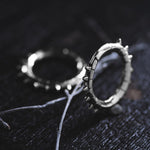 Dark-themed photograph featuring KALASH Medium Spike Hoop Clicker Ear Weights in Silver Brass by Kaya Movement, exuding a tribal punk industrial jewelry vibe. One ear weight lies flat on a dark surface while the other stands upright, accompanied by dry, thin twigs that add a contrasting natural element.