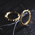 Close-up of two KALASH Medium Spike Hoop Clicker Ear Weights in Gold Brass by Kaya Movement, featuring cyborg-like designs on a dark, textured surface. One ear weight stands upright while the other lies flat. A thin, dried twig positioned diagonally beneath adds a natural element to the composition.