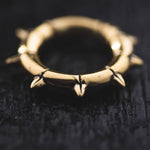 Close-up image of the KALASH Small Spike Hoop Clicker Ear Weights in Gold Brass by Kaya Movement, featuring spike-like protrusions that resemble a crown of thorns. Placed on a dark, textured surface, its metallic sheen and unique design details evoke the rugged elegance often seen in post-apocalyptic body jewelry.