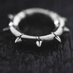Close-up of a KALASH Small Spike Hoop Clicker Ear Weights in Silver Brass by Kaya Movement, featuring thorn-like protrusions evenly spaced around its outer edge, resembling a biomechanical hoop clicker, resting on a dark, textured surface. The piece could easily be mistaken for unisex 4 gauge earrings.