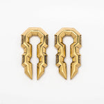 Introducing the TECHRE Futuristic Sci Fi Hexagon Keyhole Ear Weights in Gold by Kaya Movement. These bold, golden ear weights feature a sleek geometric design with sharp edges and angular shapes that create an elongated, symmetrical pattern, reminiscent of biomechanical cyborg hangers. The 0-gauge earrings are showcased against a plain white background.