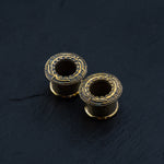 Close-up shot of two Kaya Movement's TERA Futuristic Sci Fi Flesh Tunnels in gold brass, featuring intricate patterns on their exposed circular tops. They are displayed on a black, textured surface, showcasing their hollow centers.