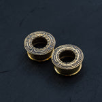 Two intricately designed TERA Futuristic Sci Fi Flesh Tunnels in gold brass from Kaya Movement, featuring a circular and ornate pattern. These futuristic ear tunnels, available in sizes ranging from 8mm to 20mm, are placed side by side on a black, textured surface, highlighting their detailed craftsmanship and metallic sheen.