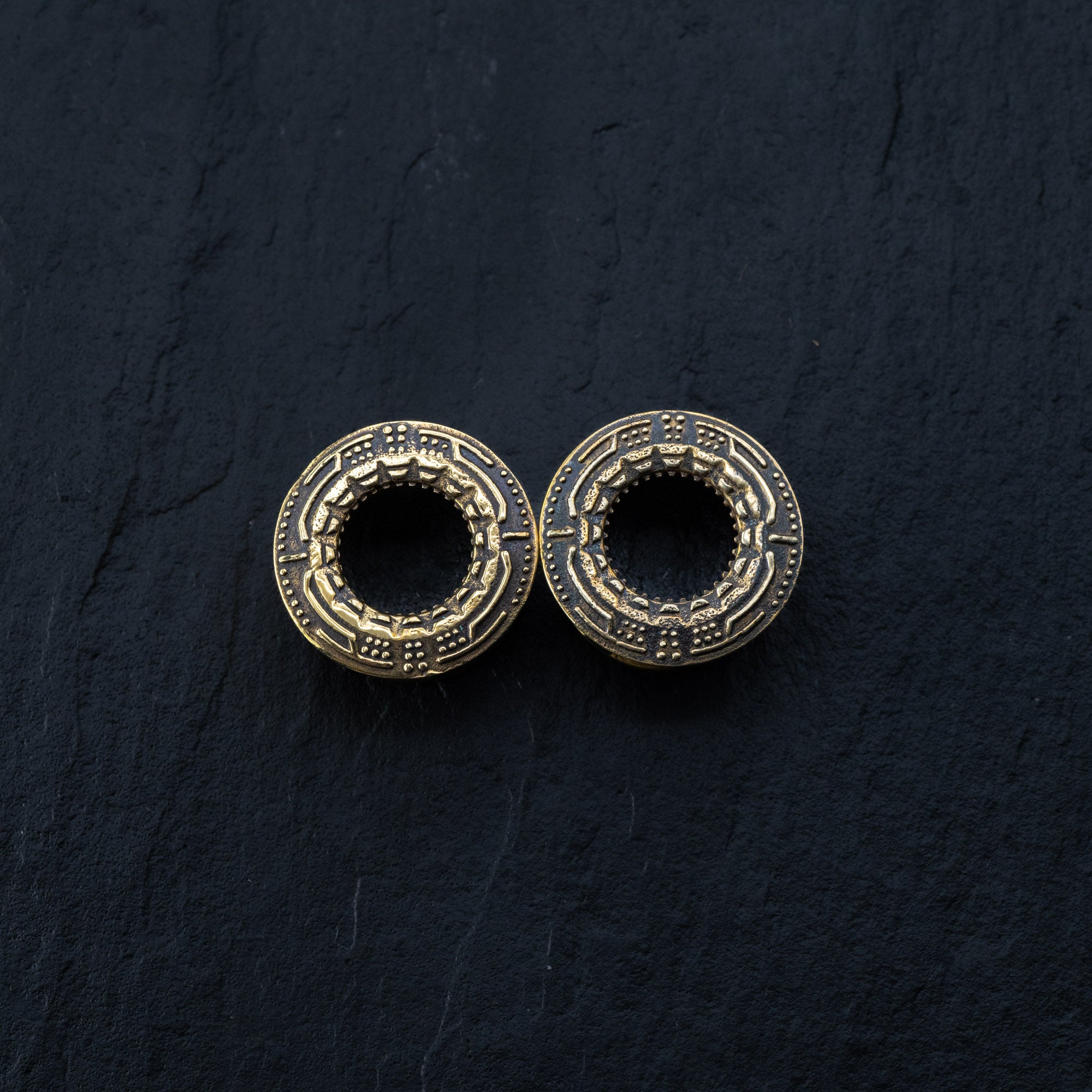 A close-up image of two TERA Futuristic Sci Fi Flesh Tunnels in gold brass by Kaya Movement, placed side by side on a dark, textured surface. These intricate pieces feature detailed patterns resembling small symbols or engravings encircling a hollow center and are available in sizes ranging from 8mm to 20mm.