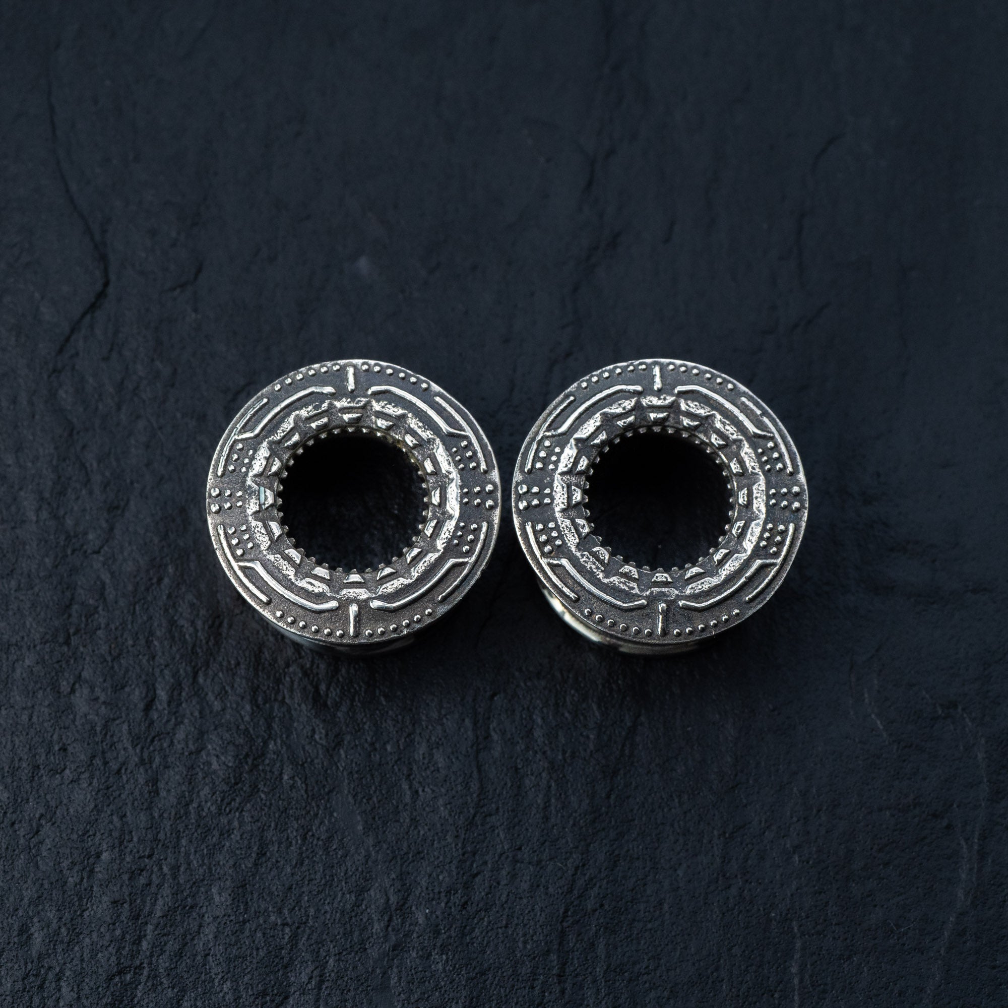 The TERA Futuristic Sci Fi Flesh Tunnels in Silver Brass from Kaya Movement are displayed on a dark, textured surface. Designed with intricate, geometric engravings around their hollow centers, these unisex ear tunnels range from 8mm to 20mm and showcase an ornate and decorative pattern—perfect for stretched ears.