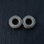 The TERA Futuristic Sci Fi Flesh Tunnels in Silver Brass from Kaya Movement are displayed on a dark, textured surface. Designed with intricate, geometric engravings around their hollow centers, these unisex ear tunnels range from 8mm to 20mm and showcase an ornate and decorative pattern—perfect for stretched ears.